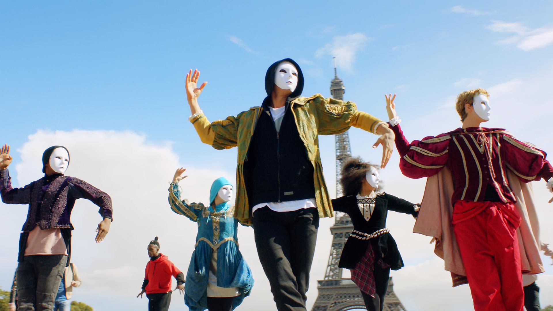 Tänzer mit weißen Masken vor dem Gesicht tanzen vor dem Eiffelturm.