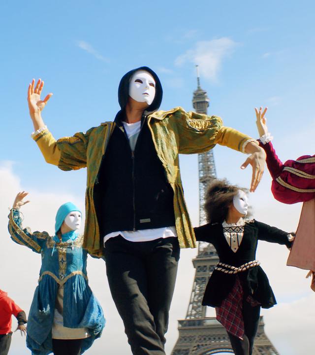 Tänzer mit weißen Masken vor dem Gesicht tanzen vor dem Eiffelturm.