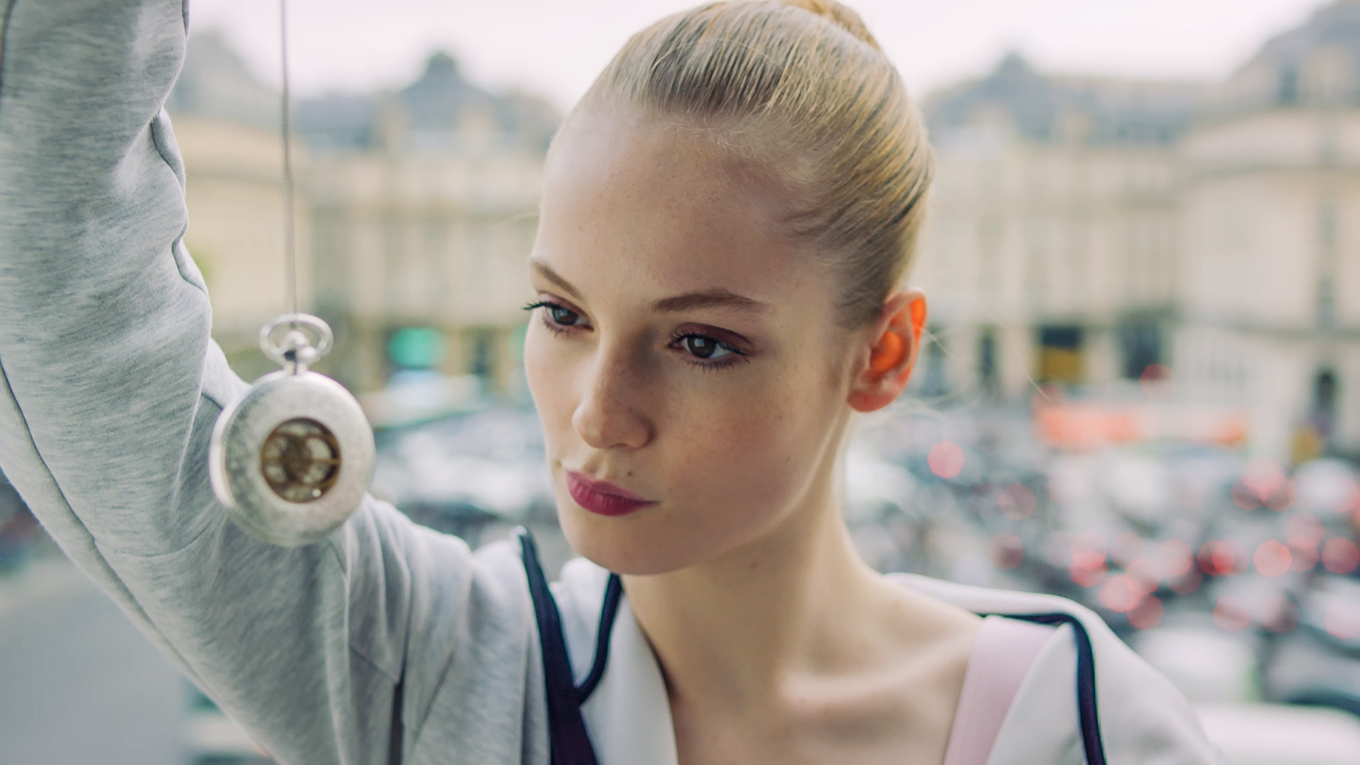 Thea (m) schaut eine silberne, ins Bild hängende Taschenuhr an, die sie an einer Schnur zum Betrachten vor ihr Gesicht hält.