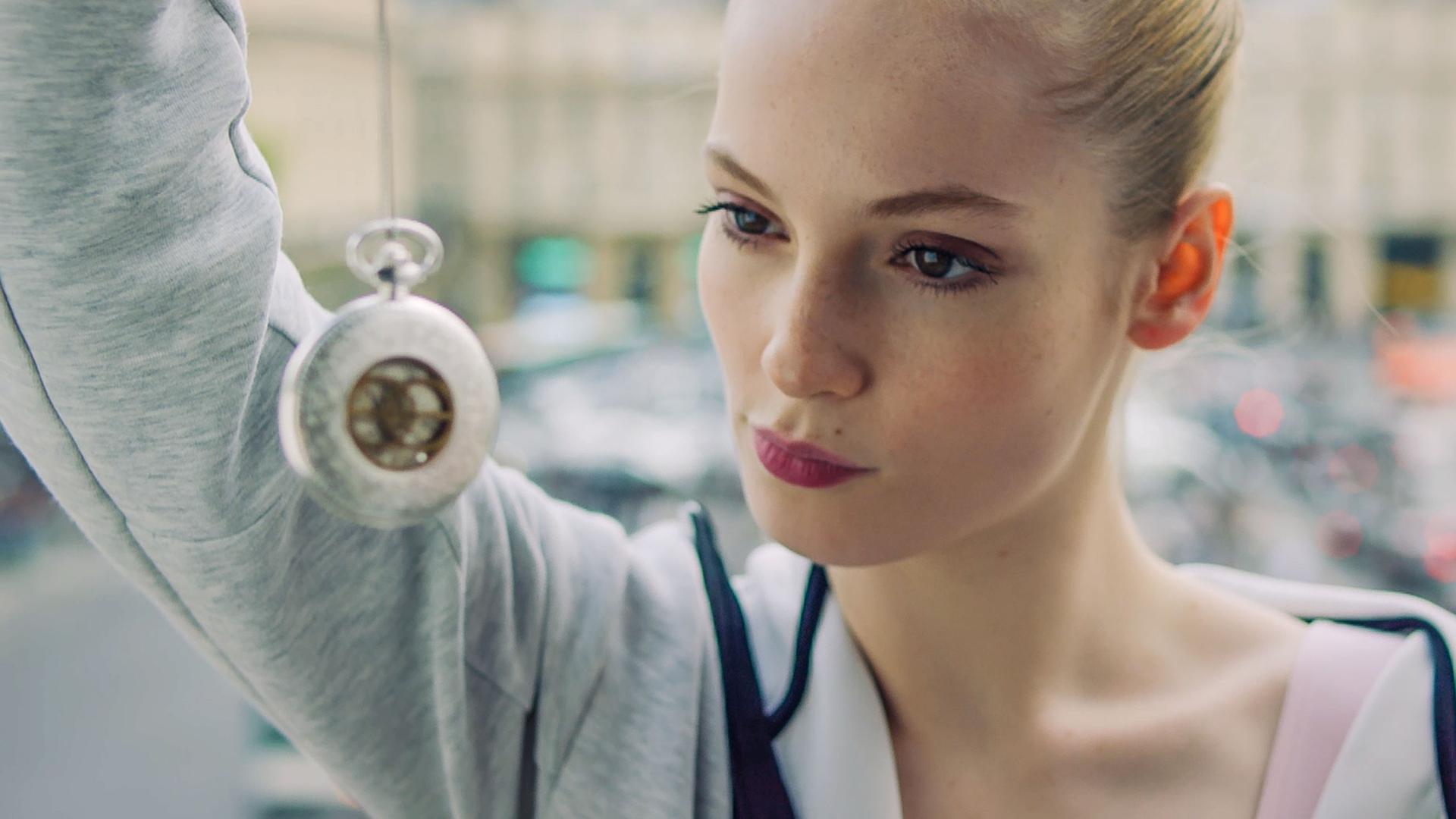 Thea schaut eine silberne, ins Bild hängende Taschenuhr an, die sie an einer Schnur zum Betrachten vor ihr Gesicht hält.