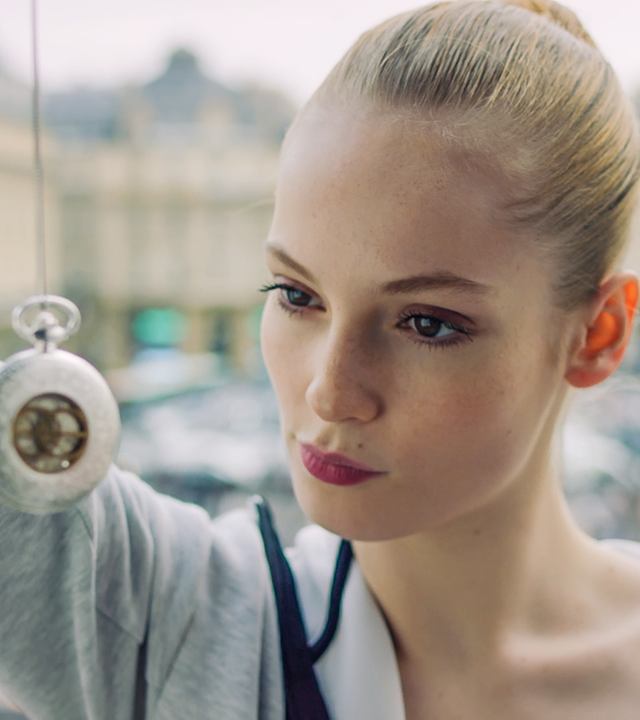 Thea (m) schaut eine silberne, ins Bild hängende Taschenuhr an, die sie an einer Schnur zum Betrachten vor ihr Gesicht hält.