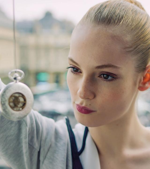 Thea schaut eine silberne, ins Bild hängende Taschenuhr an, die sie an einer Schnur zum Betrachten vor ihr Gesicht hält.