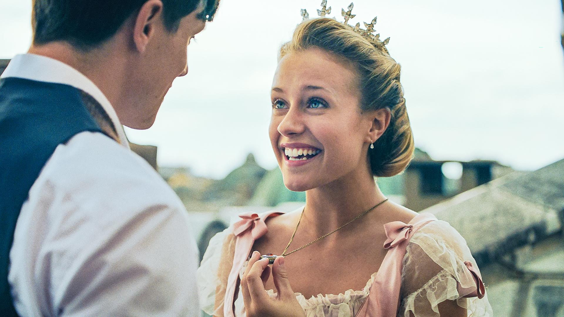 Lena steht vor einem jungen Mann und lächelt ihn an. Sie trägt eine Krone.