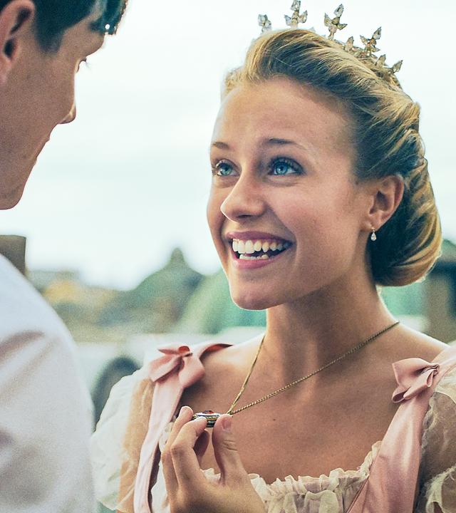 Lena steht vor einem jungen Mann und lächelt ihn an. Sie trägt eine Krone.