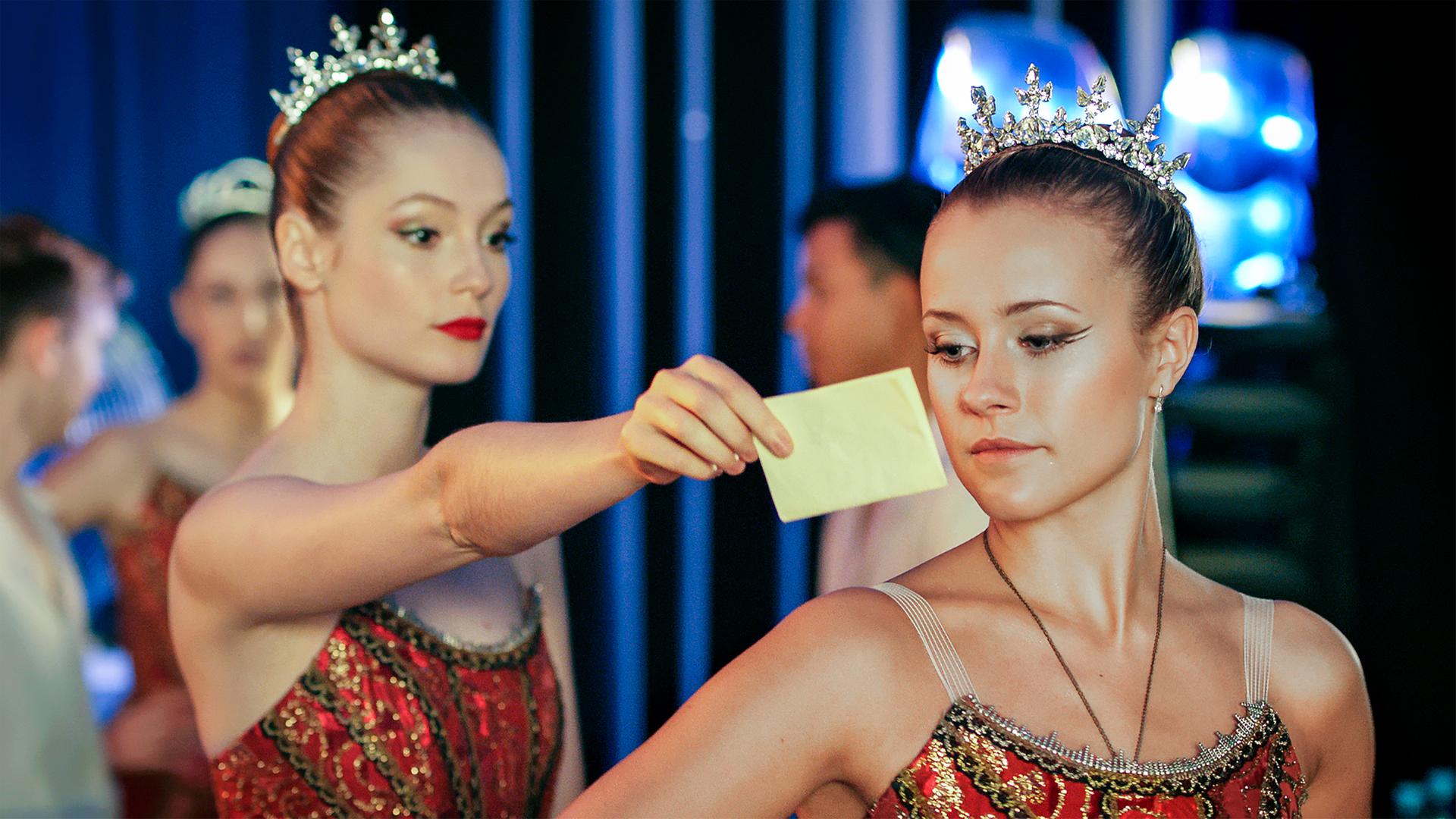 Thea zeigt Lena einen Brief. Sie haben beide sehr schöne feierliche Kleider und eine kleine Krone an.