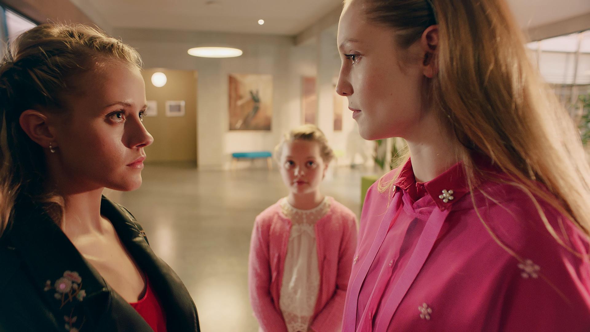 Lena steht vor einer jungen Frau. Sie schauen einander mit einem durchdringenden Blick an. Im Hintergrund steht ein kleines Mädchen und sieht die beiden an.