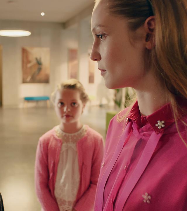 Lena steht vor einer jungen Frau. Sie schauen einander mit einem durchdringenden Blick an. Im Hintergrund steht ein kleines Mädchen und sieht die beiden an.