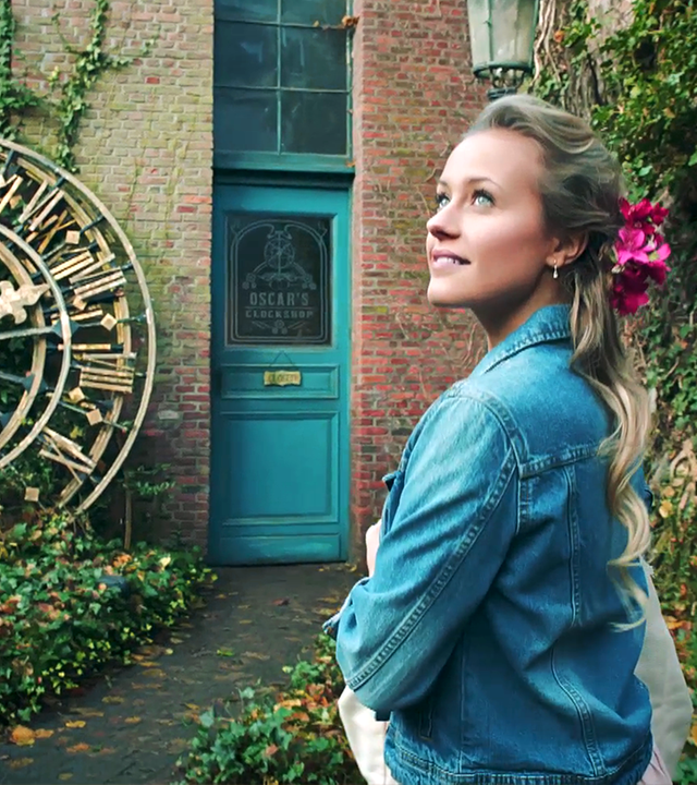 Lena blickt in den Himmel. Hinter ihr lehnt eine große goldene Uhr an einer Hauswand.