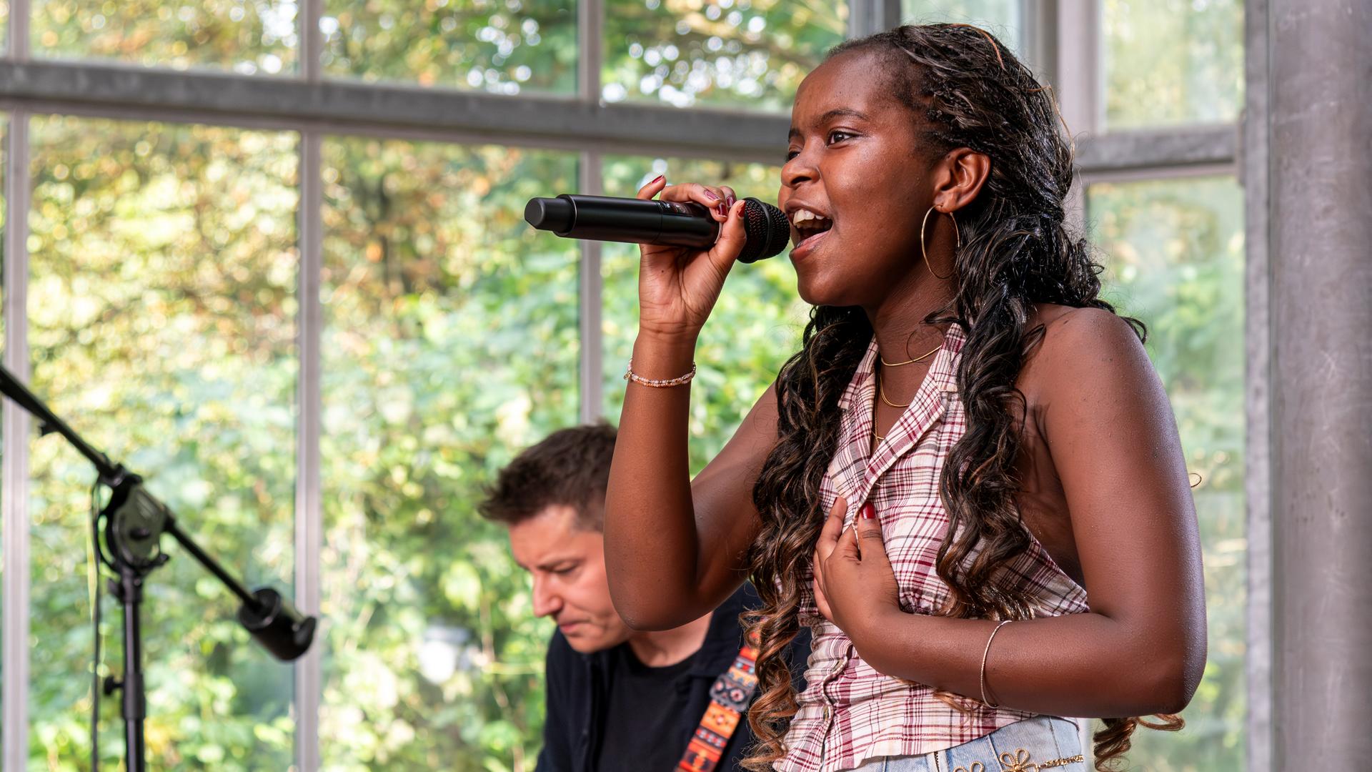 Das Bild zeigt die Kandidatin Asmina, die ein schwarzes Mikrofon in ihrer Hand hält. Im Hintergrund ist der Gitarrist der "Dein Song"-Band zu erkennen. Asmina steht in einem alten Gewächshaus.