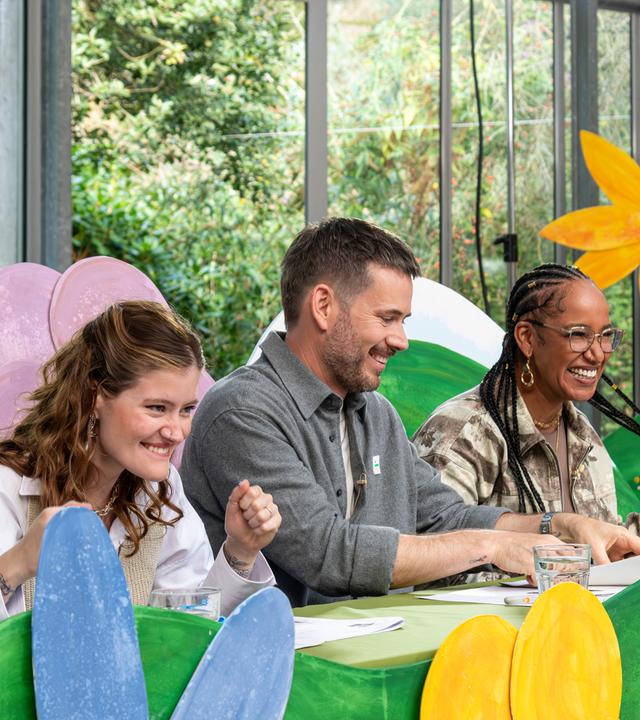 "Dein Song - Entscheidung im Halbfinale":  Die Dein Song Jury um Madeline Juno, Johannes Strate und Aisha Vibes (v.l.n.r.) sitzt lachend nebeneinander an einem Tisch inmitten einer bunten Blumenkulisse.