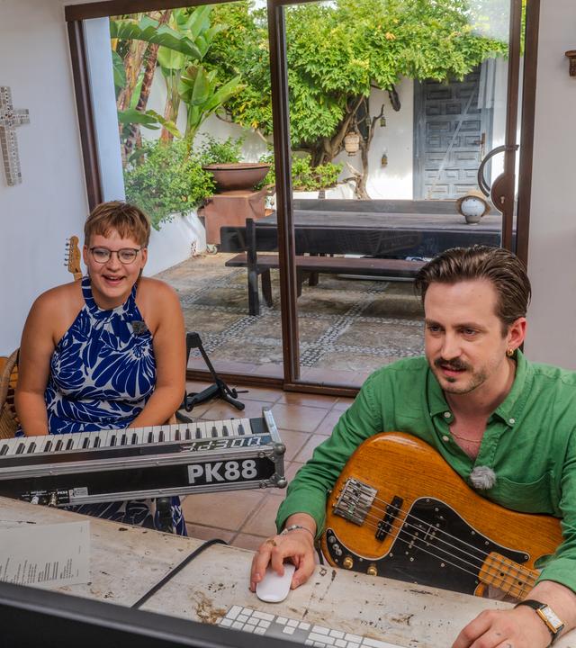 Das Bild zeigt die "Dein Song"-Kandidatin Matilda mit ihrem Paten Marti Fischer (v.l.n.r.). Matilda sitz an einem E-Piano und Marti bedient mit seiner rechten Hand eine Maus während er eine E-Gitarre auf dem Schoß hat. Die beiden sitzen in einer Finca auf Ibiza.