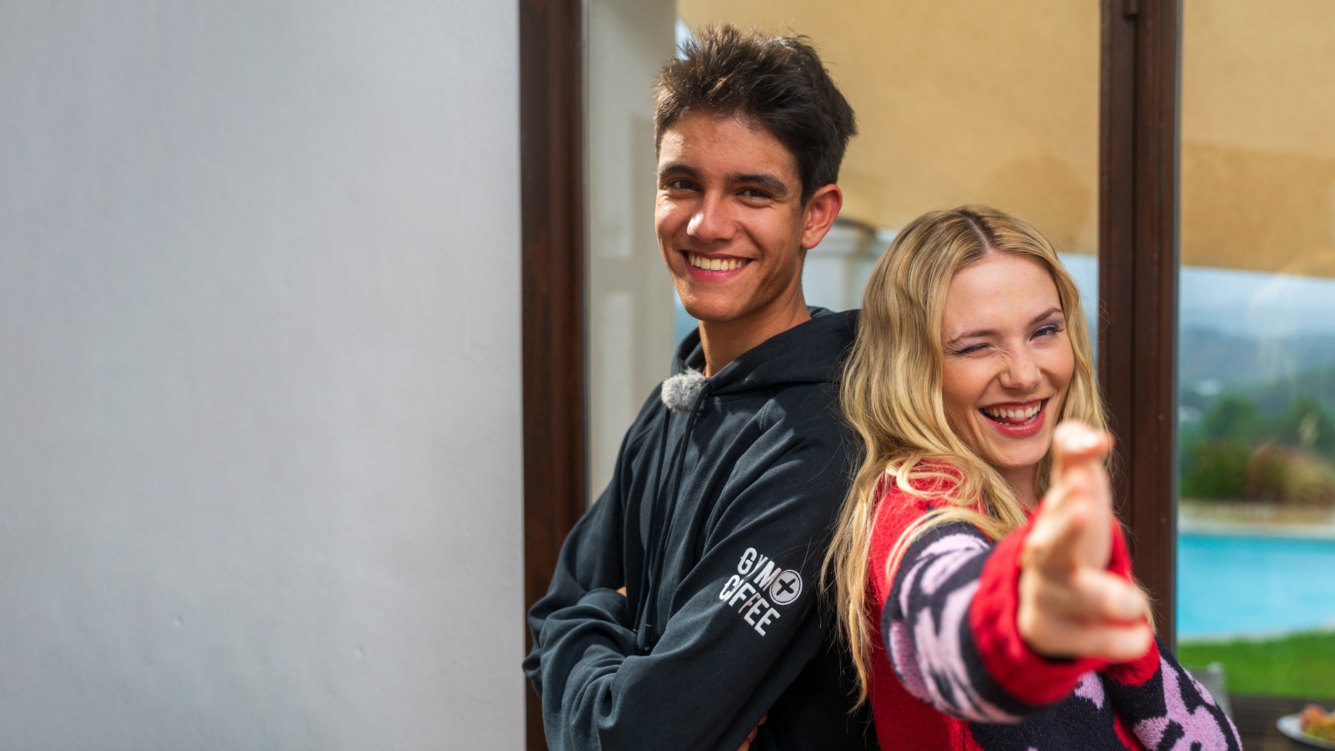 Links im Bild ist der "Dein Song"-Kandidat Luis zu sehen. Er lächelt in die Kamera und steht Rücken an Rücken mit seiner Patin Esther Graf, die ebenfalls in die grinst und zwei Finger in die Kamera streckt.