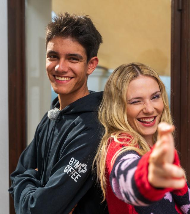 Links im Bild ist der "Dein Song"-Kandidat Luis zu sehen. Er lächelt in die Kamera und steht Rücken an Rücken mit seiner Patin Esther Graf, die ebenfalls in die grinst und zwei Finger in die Kamera streckt.