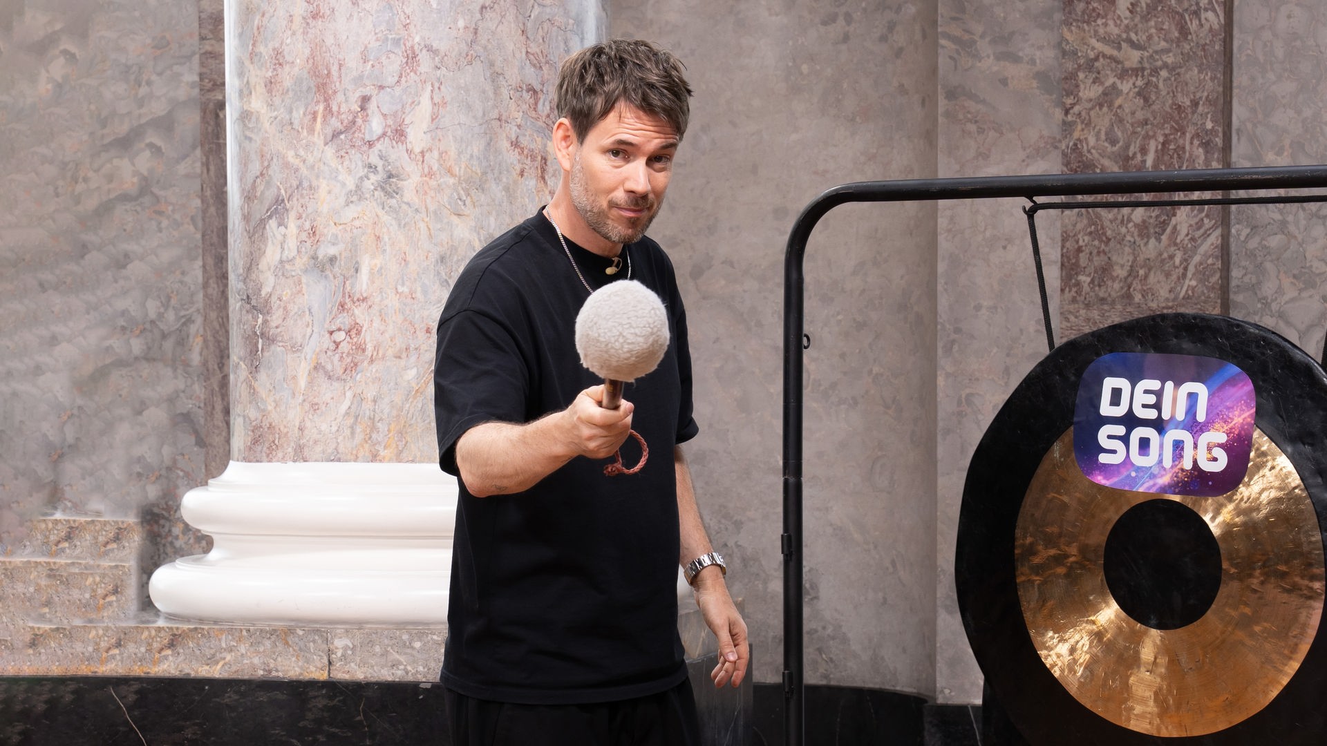 "Dein Song - Gong, Gong, Gong": Johannes Strate hält einen Schlägel in der Hand und steht links von einem Gong, auf dem das Dein Song Logo zu sehen ist. Während er in die Kamera guckt holt er zum Schlag aus. Im Hintergrund ist die Rotunde von Schloss Biebrich zu erkennen.
