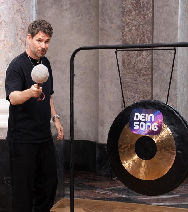 "Dein Song - Gong, Gong, Gong": Johannes Strate hält einen Schlägel in der Hand und steht links von einem Gong, auf dem das Dein Song Logo zu sehen ist. Während er in die Kamera guckt holt er zum Schlag aus. Im Hintergrund ist die Rotunde von Schloss Biebrich zu erkennen.