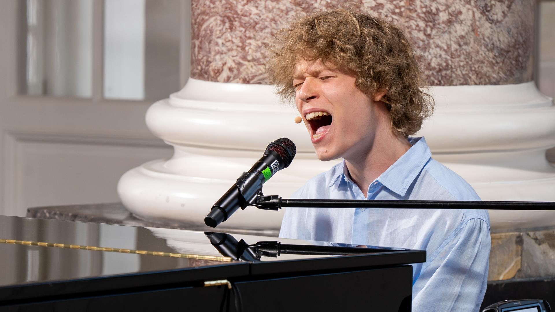 "Dein Song - Das Camp ruft!": "Dein Song" Kandidat Leander sitzt an einem Konzertflügel in Schloss Biebrich. Er singt mit weit geöffnetem Mund in ein Mikrofon, dass an einem schwarzen Ständer befestigt ist.