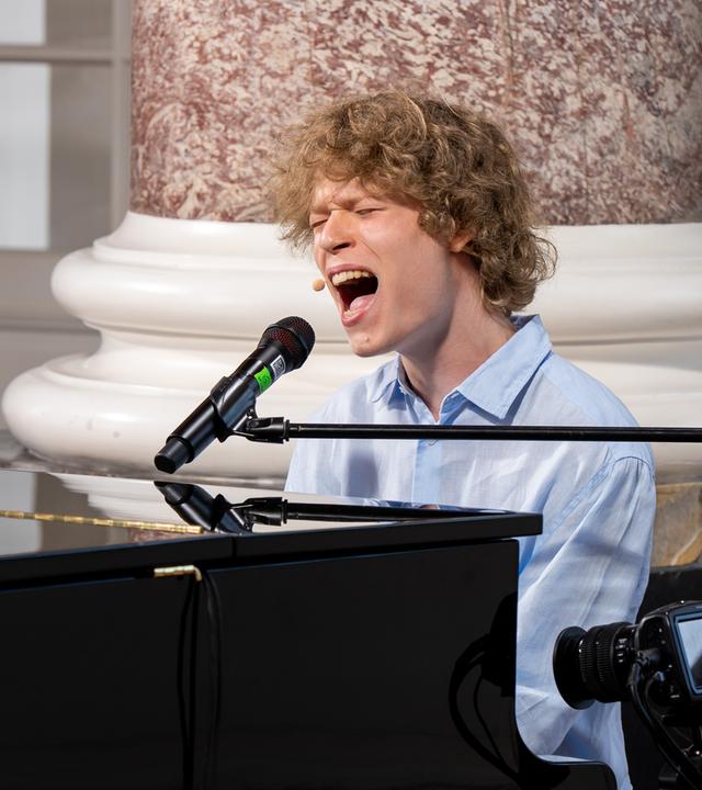 "Dein Song - Das Camp ruft!": "Dein Song" Kandidat Leander sitzt an einem Konzertflügel in Schloss Biebrich. Er singt mit weit geöffnetem Mund in ein Mikrofon, dass an einem schwarzen Ständer befestigt ist.