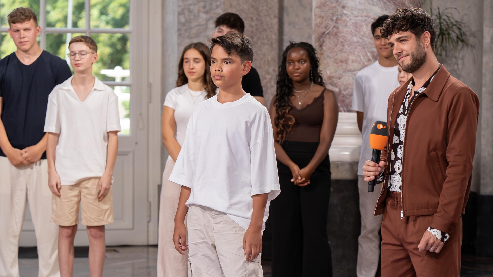"Dein Song - Der Moment der Entscheidung": Rechts im Bild steht "Dein Song" Moderator Luca Hänni mit einem ZDF Mikrofon in der Hand. Rechts von ihm steht der Kandidat Samuel. Die zwei stehen in der Mitte der Rotunde von Schloss Biebrich. Im Hintergrund sind sechs weitere Kandis zu sehen. Alle blicken in die linke Bildhälfte.