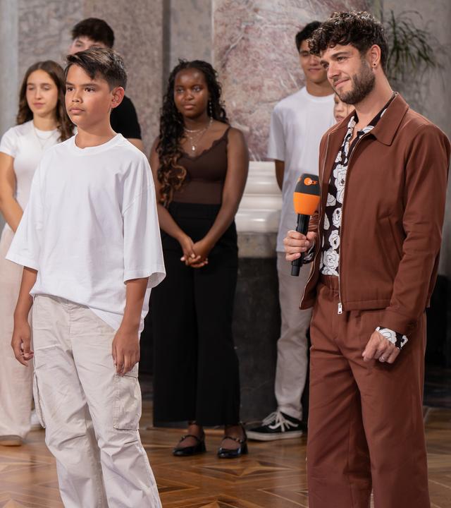 "Dein Song - Der Moment der Entscheidung": Rechts im Bild steht "Dein Song" Moderator Luca Hänni mit einem ZDF Mikrofon in der Hand. Rechts von ihm steht der Kandidat Samuel. Die zwei stehen in der Mitte der Rotunde von Schloss Biebrich. Im Hintergrund sind sechs weitere Kandis zu sehen. Alle blicken in die linke Bildhälfte.