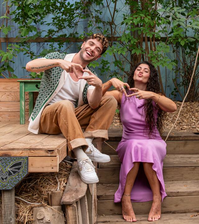 "Dein Song - Campstart!": Das Dein Song Moderationsduo Luca Hänni und Jeannie Wagner sitzen nebeneinander, Luca links auf einer Holzbühne, Jeannie rechts neben ihm auf einer Holztreppe. Beide machen eine Herzgeste und lächeln in die Kamera.