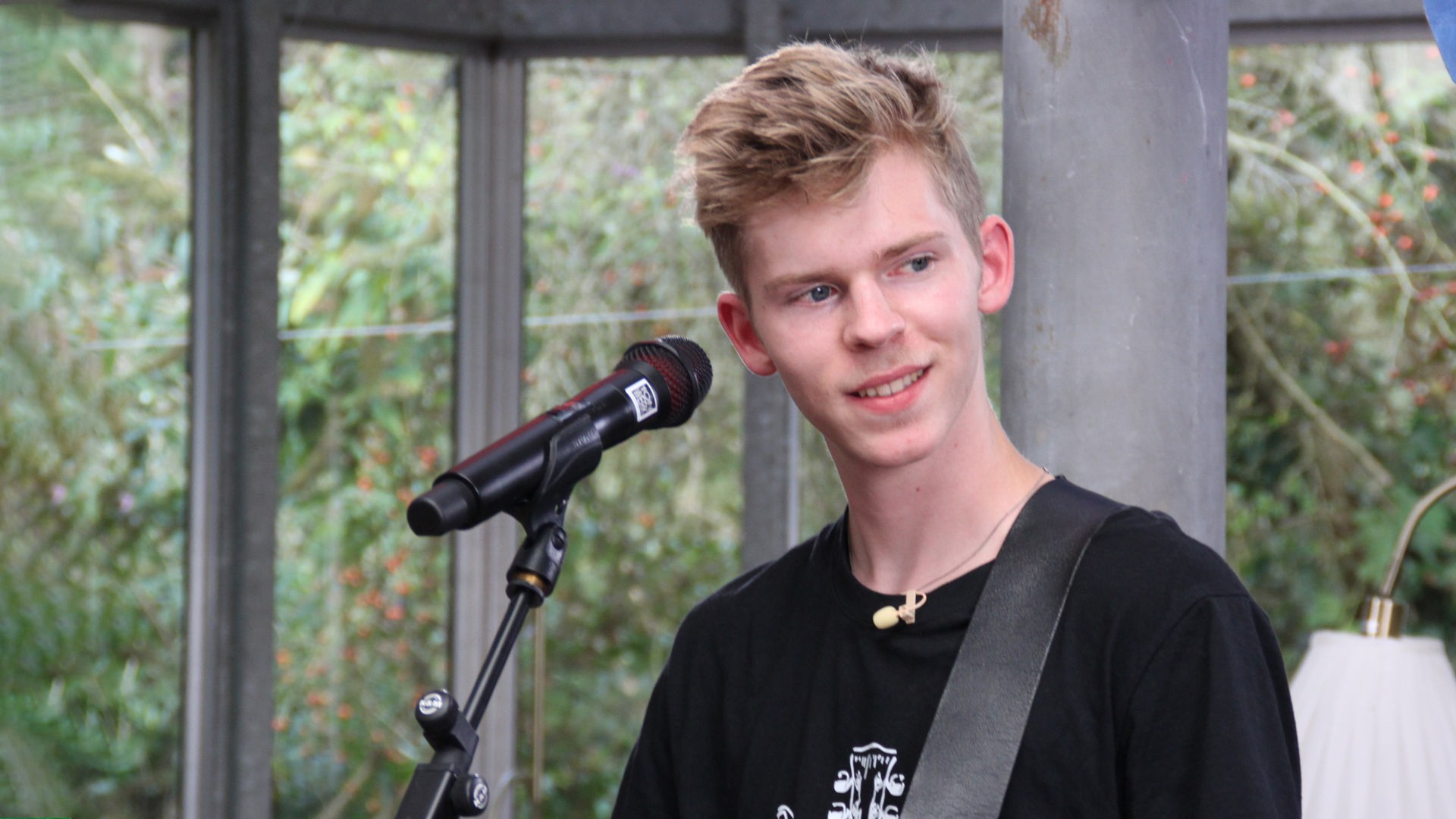 "Dein Song - Mut zur Veränderung": Dein Song Kandidat Marvin steht lächelnd vor einem Mikrofonständer, blickt lächelnd zum linken Bildrand und hat eine E-Gitarre umgeschnallt. Rechts neben ihm sitzt die Sängerin der Dein Song Band auf einem Hocker und bereitet sich vor.