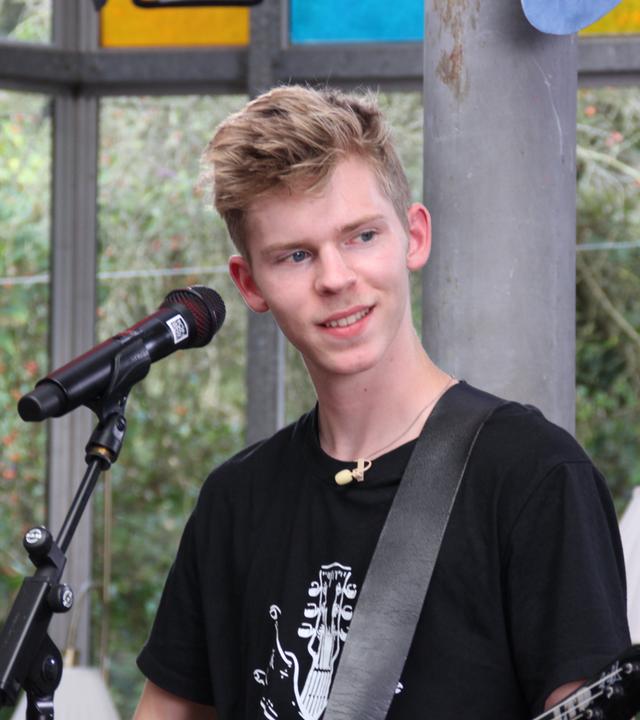 "Dein Song - Mut zur Veränderung": Dein Song Kandidat Marvin steht lächelnd vor einem Mikrofonständer, blickt lächelnd zum linken Bildrand und hat eine E-Gitarre umgeschnallt. Rechts neben ihm sitzt die Sängerin der Dein Song Band auf einem Hocker und bereitet sich vor.