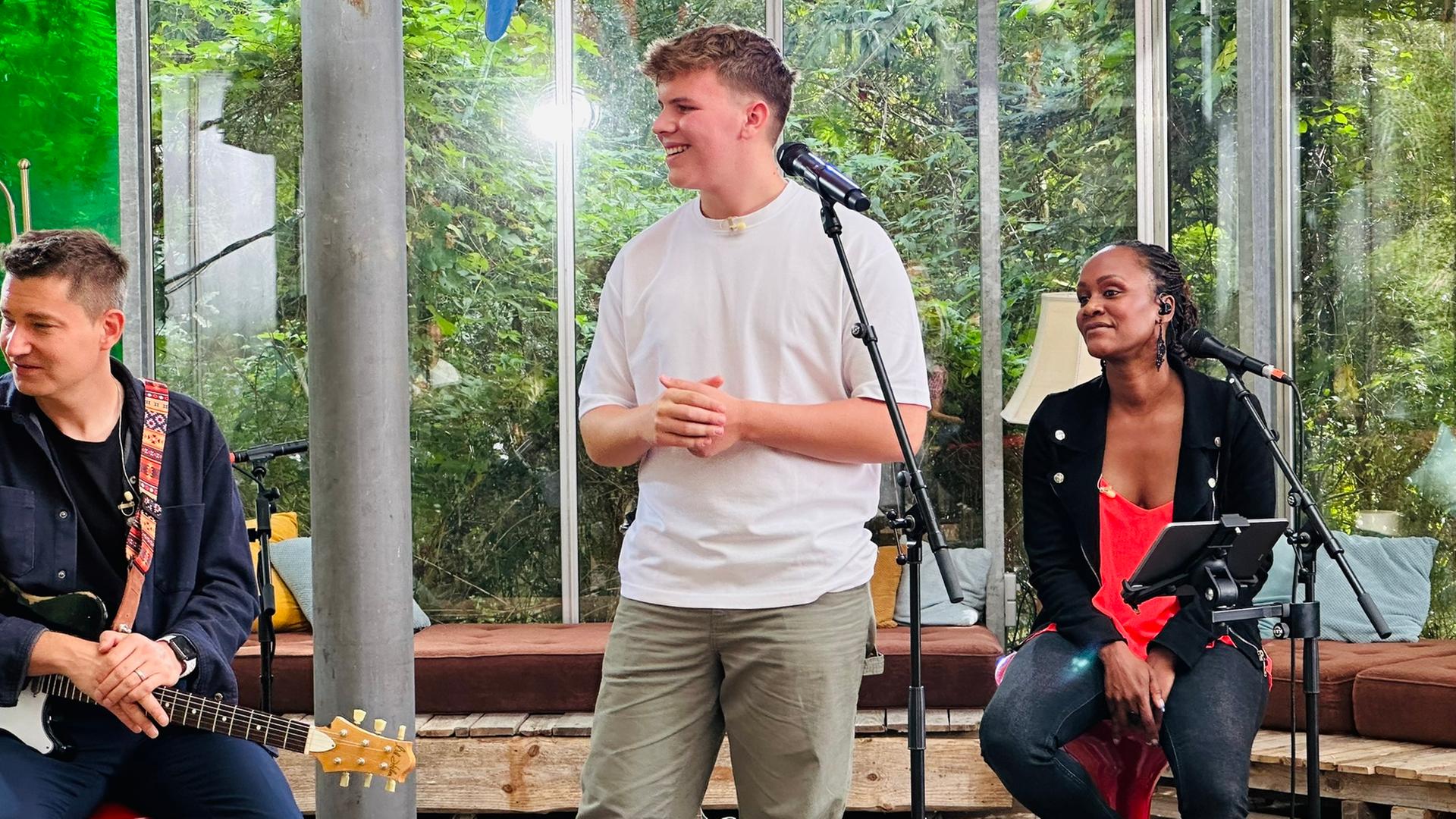 Links im Bild ist "Dein Song"-Kandidat Tom, der in die linke Bildhälfte blickt. Die Sängerin Sherita, aus der "Dein Song"-Band, sitz in der rechten Bildhälte auf einem Hocker und sieht ebenfalls zur linken Seite. Die Beiden befinden sich in einem großen alten Gewächshaus. Beide haben jeweils ein Mikrofon vor sich stehen.
