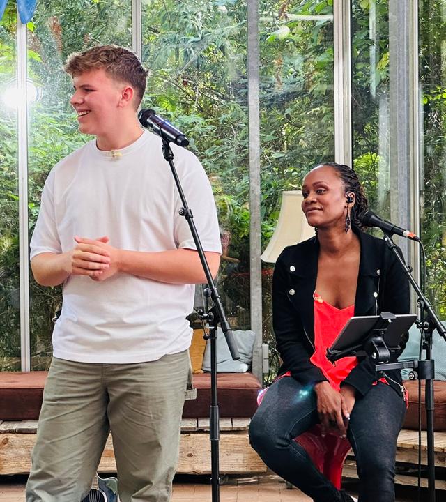 Links im Bild ist "Dein Song"-Kandidat Tom, der in die linke Bildhälfte blickt. Die Sängerin Sherita, aus der "Dein Song"-Band, sitz in der rechten Bildhälte auf einem Hocker und sieht ebenfalls zur linken Seite. Die Beiden befinden sich in einem großen alten Gewächshaus. Beide haben jeweils ein Mikrofon vor sich stehen.