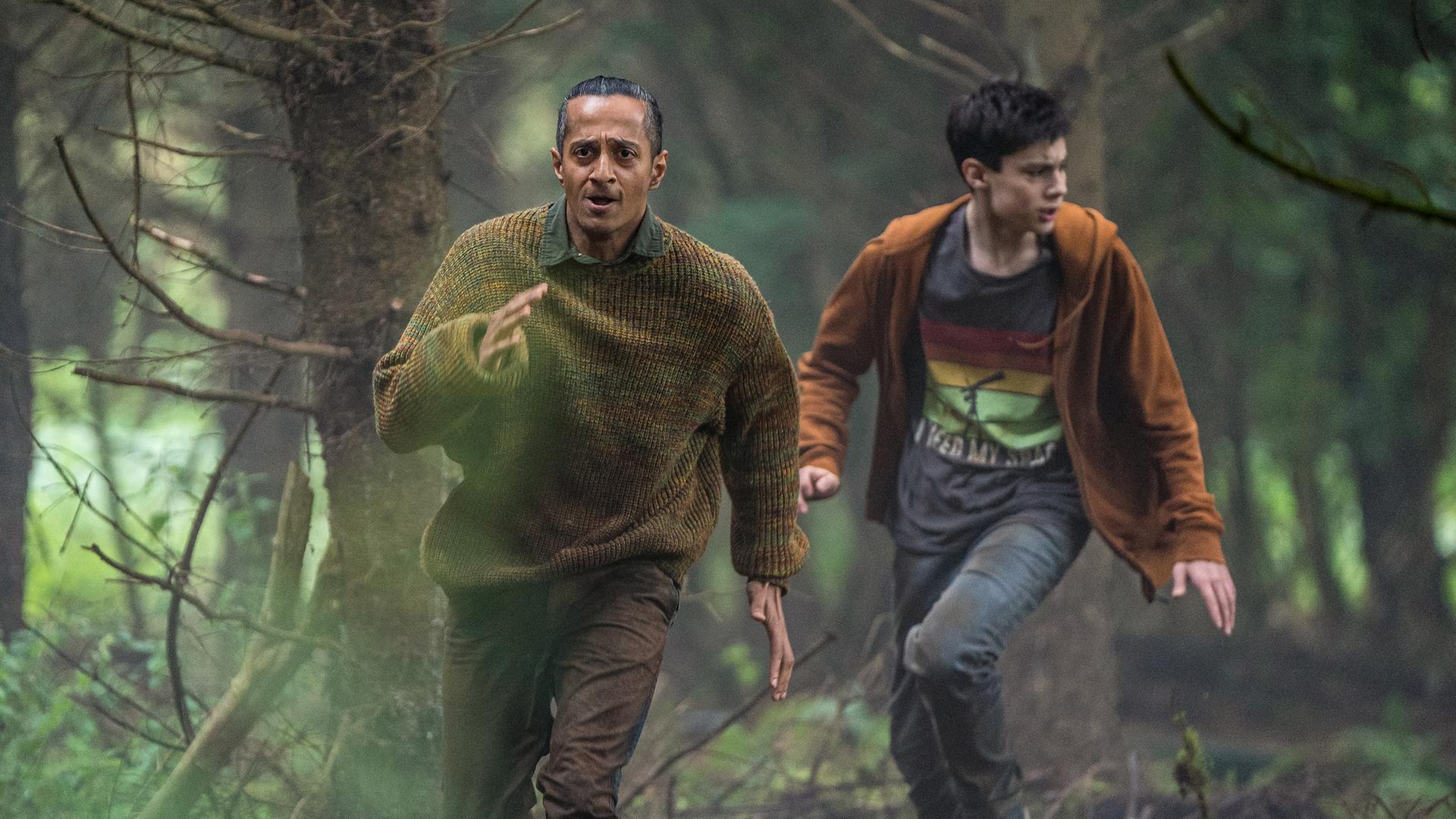Louis und Fischer laufen bei Tageslicht durch den Wald, der durch einen Grünfilter besonders grün aussieht. Sie sind in Aufruhr mit ängstlich verzerrten Gesichtern.