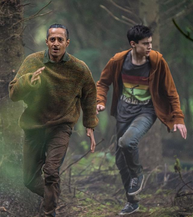 Louis und Fischer laufen bei Tageslicht durch den Wald, der durch einen Grünfilter besonders grün aussieht. Sie sind in Aufruhr mit ängstlich verzerrten Gesichtern.