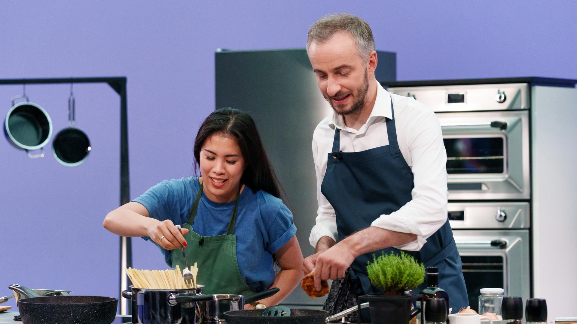 Böhmi brutzelt - Folge 04 mit Mai Thi Nguyen-Kim und Jan Böhmermann, der Kochtalk bei ZDFneo. Beide stehen in Schürze hinter einem Herd.