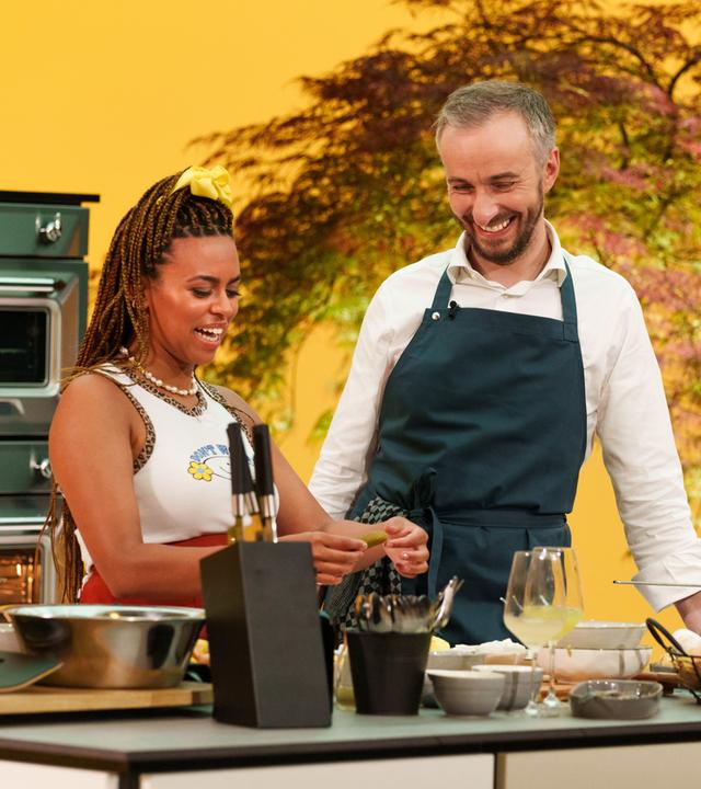 Aminata Belli und Jan Böhmermann stehen nebeneinander lachend in einer Küche.