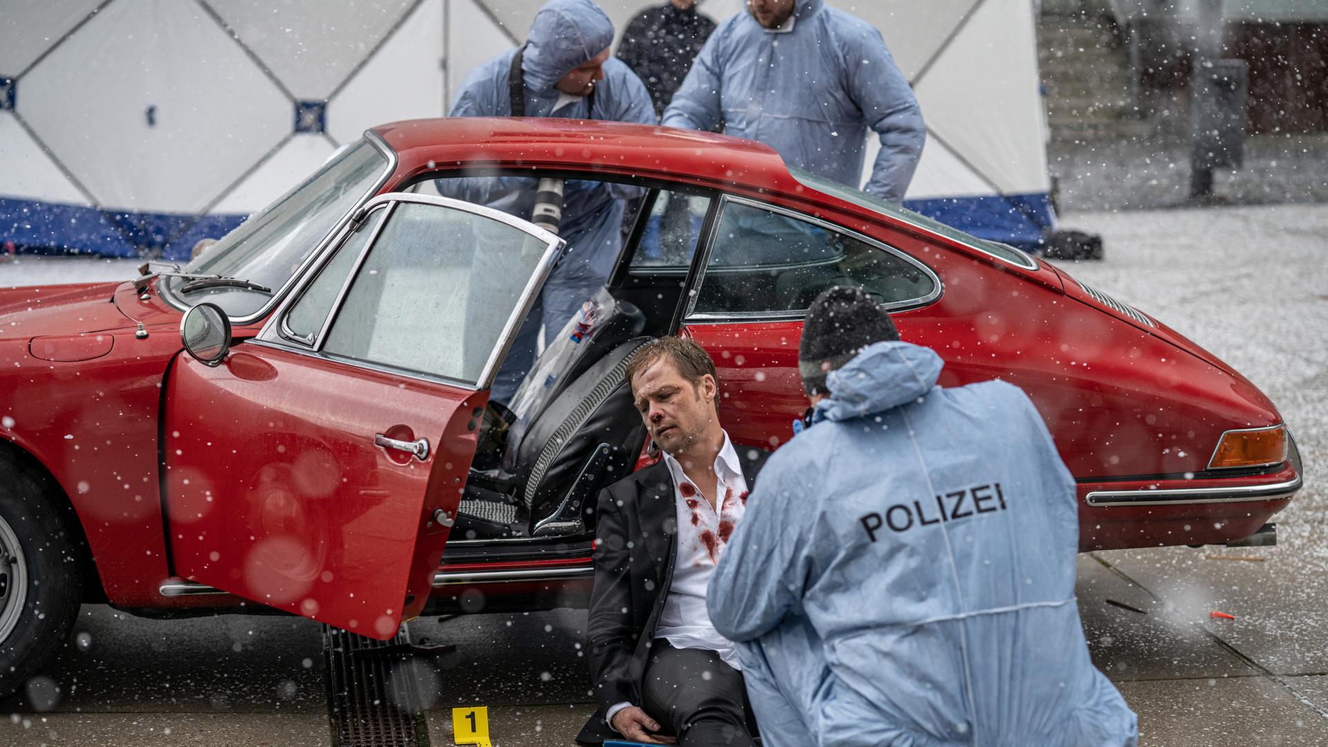 Man sieht einen Polizisten und einen blutenden Mann vor einem roten Auto.