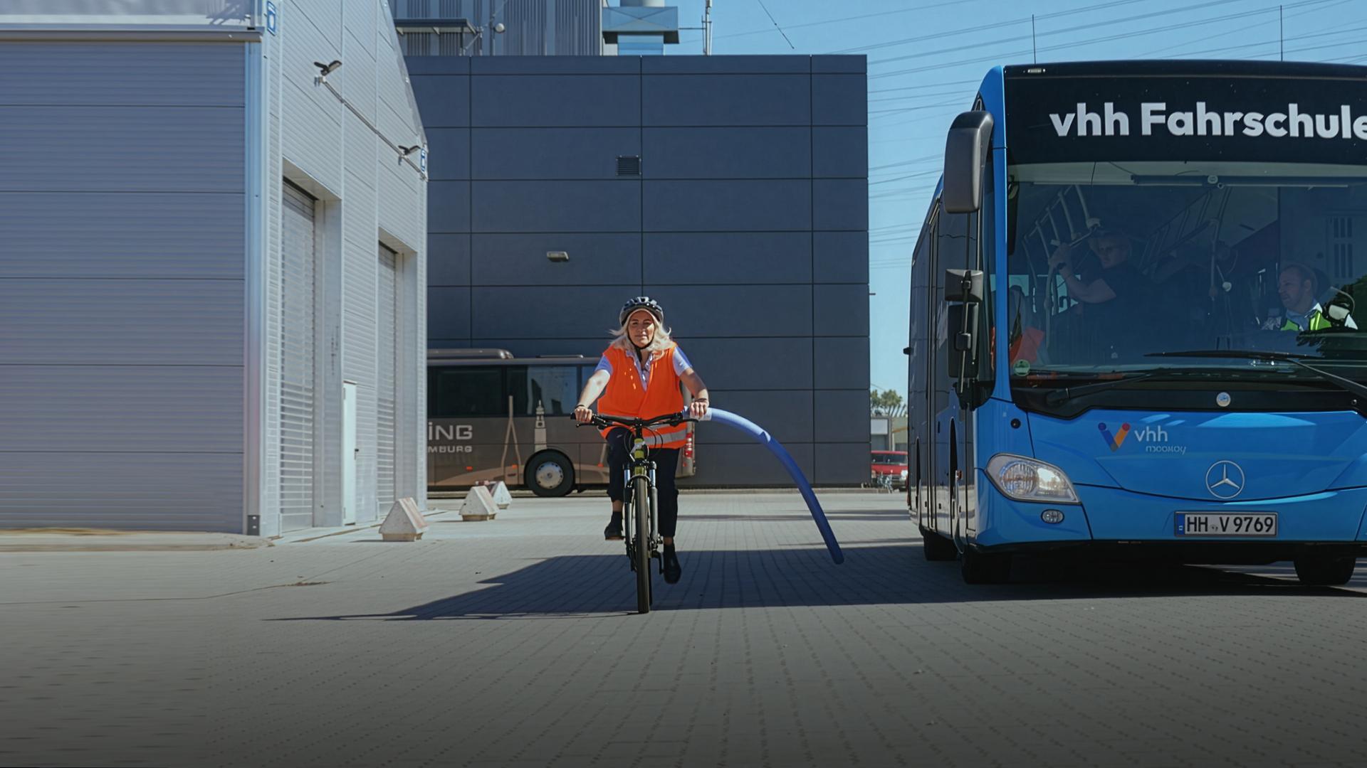 Eine Frau fährt mit Helm und Warnweste auf einem Fahrrad neben einem Bus her. Am Lenkrad des Rads ist eine Schwimmnudel befestigt. Diese zeigt den Sicherheitsabstand von 1,50 Metern an. Auf dem blauen Bus steht oben der Schriftzug „vhh Fahrschule“.