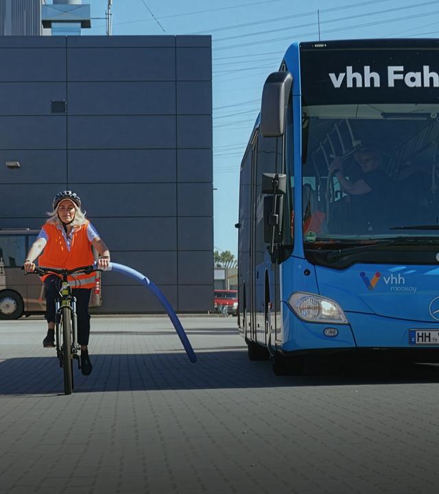 Eine Frau fährt mit Helm und Warnweste auf einem Fahrrad neben einem Bus her. Am Lenkrad des Rads ist eine Schwimmnudel befestigt. Diese zeigt den Sicherheitsabstand von 1,50 Metern an. Auf dem blauen Bus steht oben der Schriftzug „vhh Fahrschule“.
