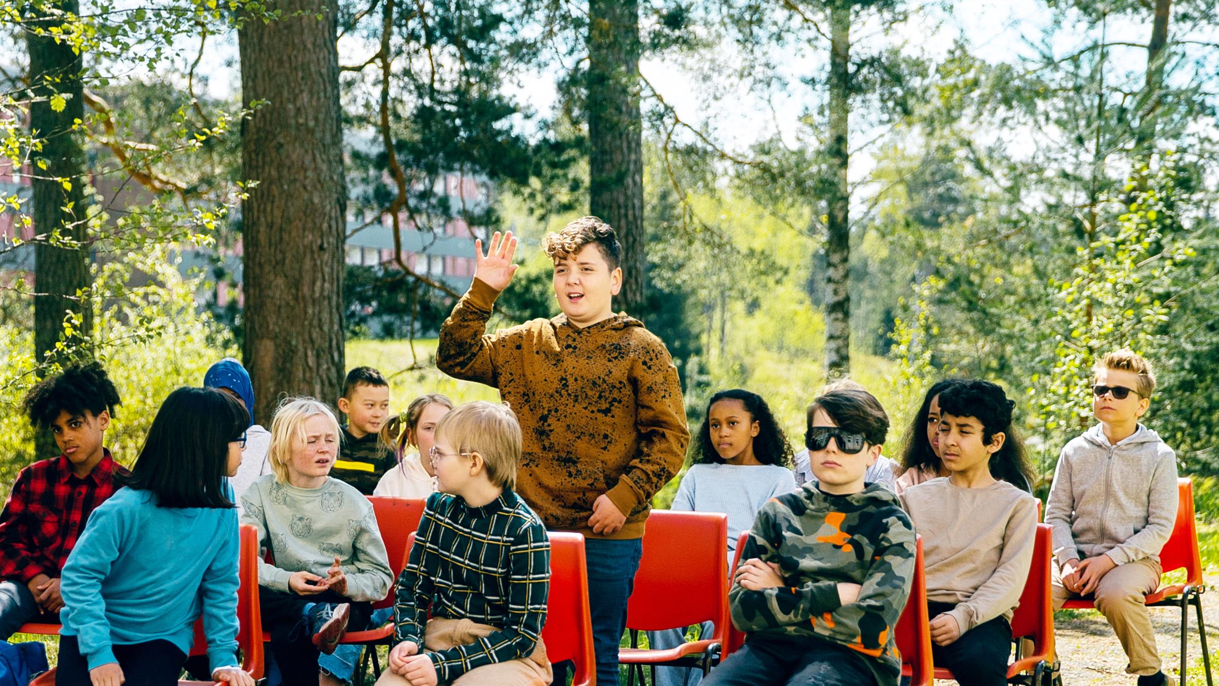 Blaze steht mit schüchternem Blick und nach oben gestreckter Hand inmitten seiner Mitschüler, die alle um ihn herum im Freien auf Stühlen sitzen und ihn gespannt anschauen.