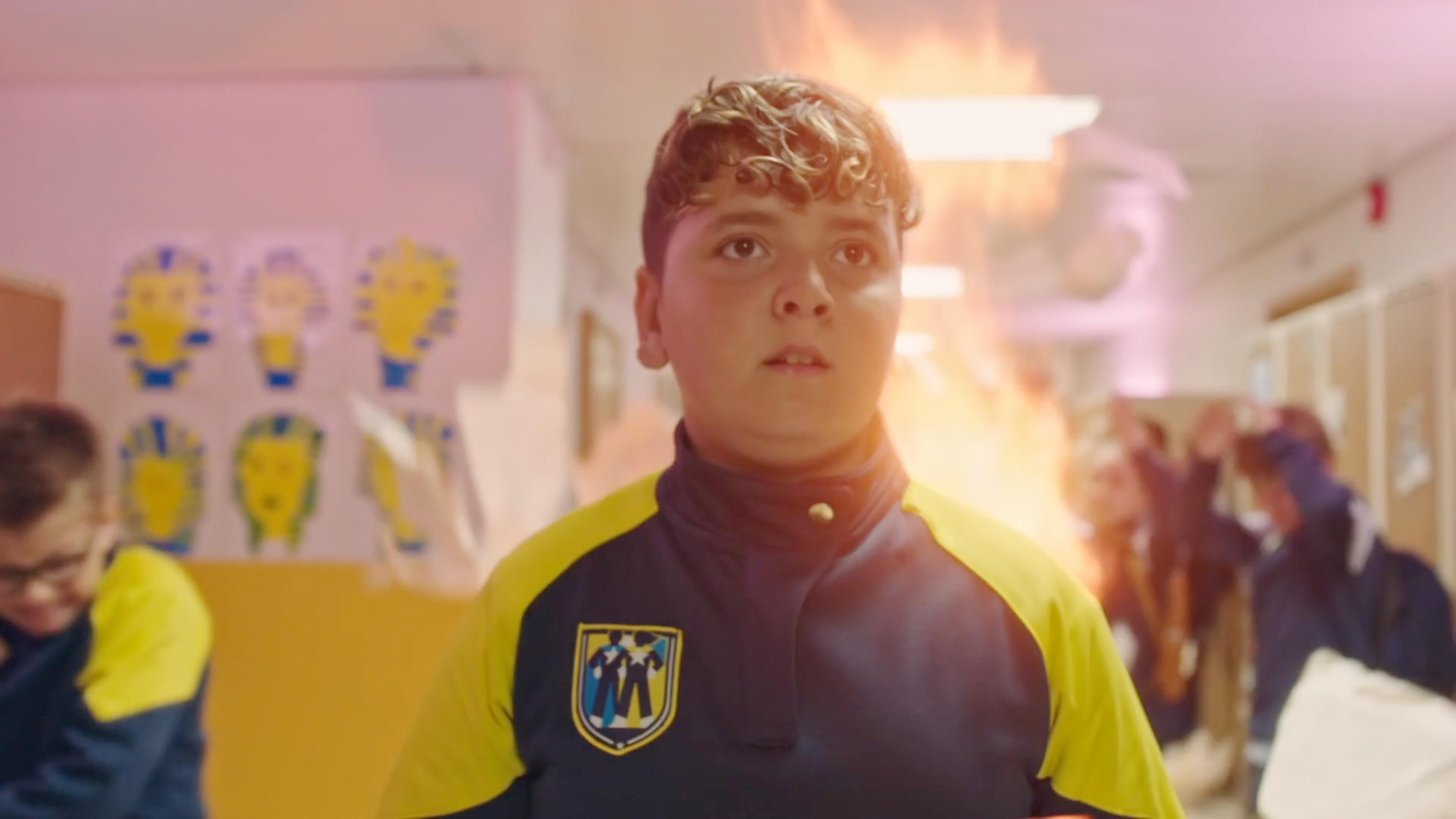 Blaze steht inmitten des Schulflures und sein Blick geht angespannt nach vorne. Hinter ihm lodert eine Stichflamme und einige Schüler rennen im Hintergrund aufgeregt durch den Flur.