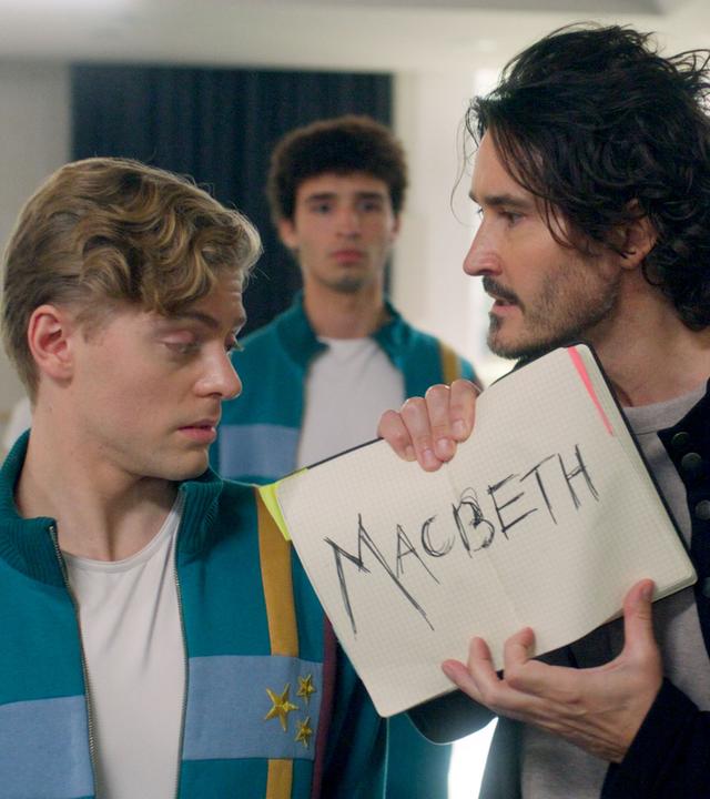 Die Schüler befinden sich im Probenraum. Finn schaut geschockt auf das aufgeschlagene Notizbuch mit der Aufschrift "Macbeth", das ihm Ballettmeister Armando unter die Nase hält.