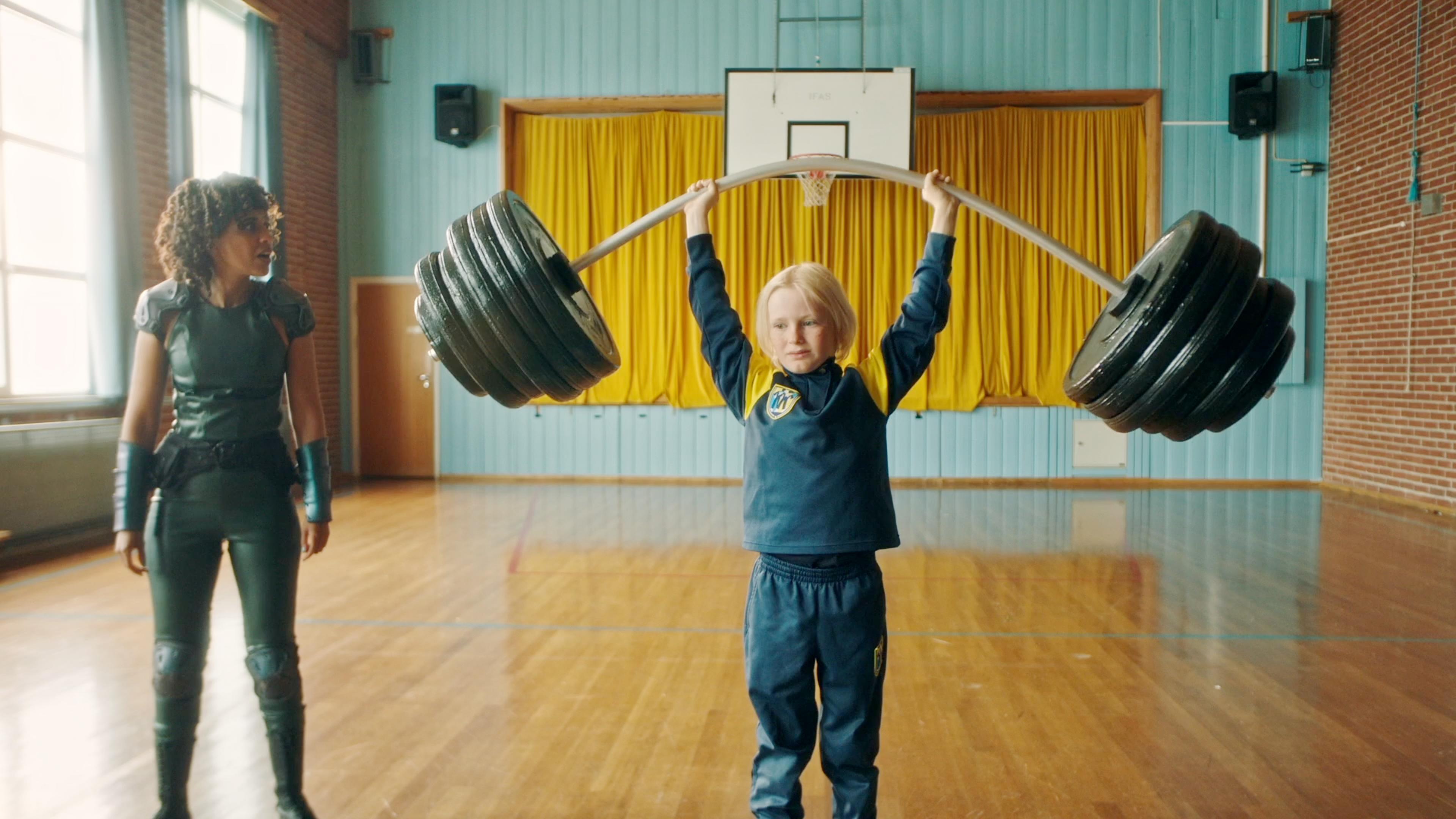 In der Schulturnhalle: Lehrerin Tundra steht mit erstauntem Blick neben Raymond, der eine riesige Hantel mit Gewichten nach oben streckt.