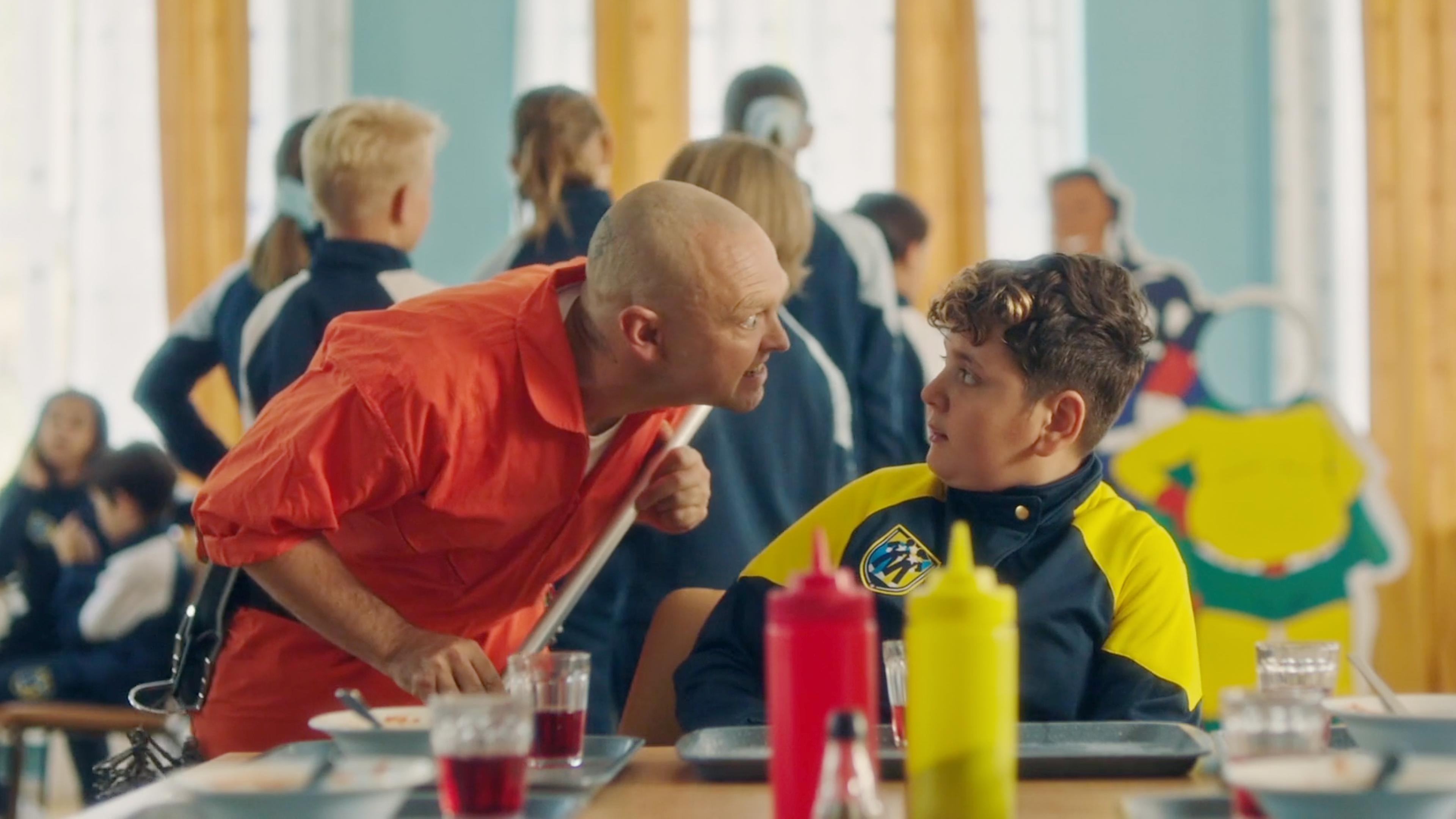 Blaze sitzt in der Schulkantine am Tisch und schaut verängstigt zu Hausmeister Joe, der neben ihm steht und sich fies blickend zu ihm herunterbeugt. Im Hintergrund sind mehrere Mitschüler:innen zu sehen.