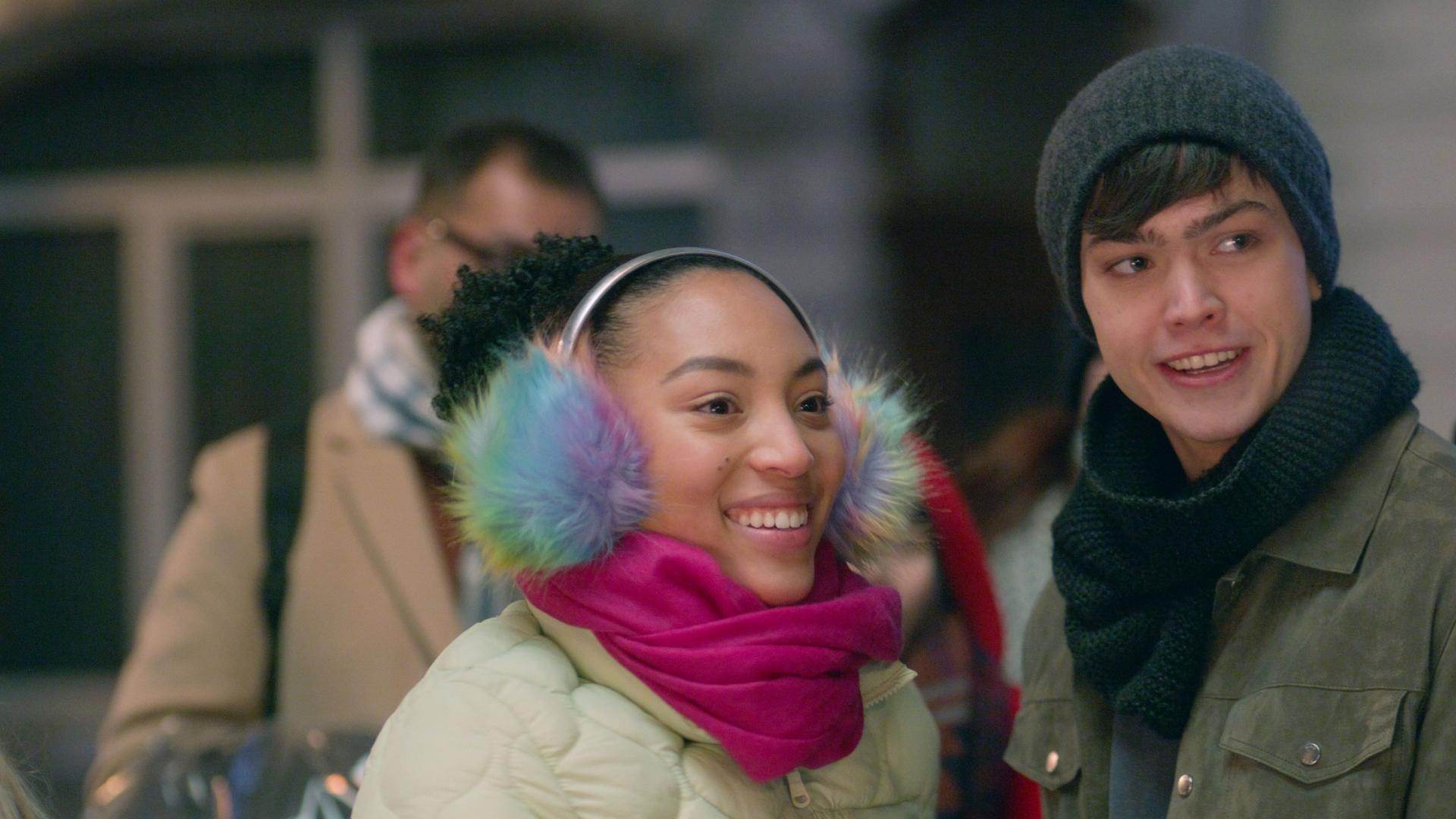 Cece trägt bunte Ohrenschützer, eine dicke Jacke und einen pinkfarbenen Schal. Sie steht glücklich lächelnd neben Adrian.