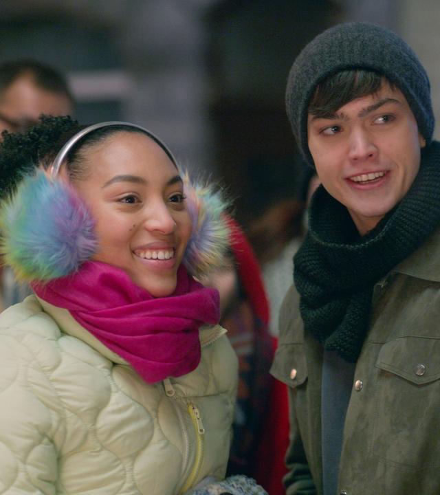 Cece trägt bunte Ohrenschützer, eine dicke Jacke und einen pinkfarbenen Schal. Sie steht glücklich lächelnd neben Adrian.