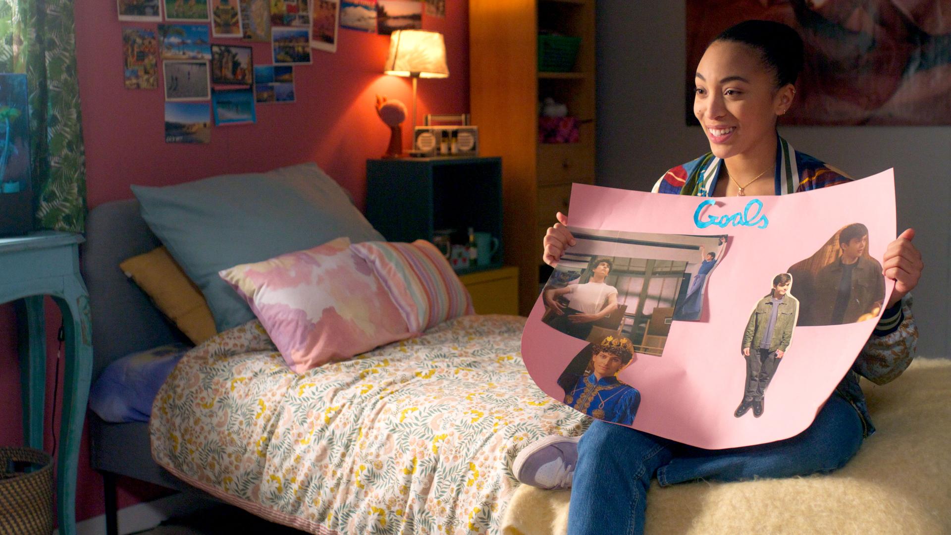 Cece sitzt lächelnd auf ihrem Bett und hält ein selbstgemachtes Plakat in die Luft, auf dem sie Bilder von Adrian und Benoit gepostet hat.