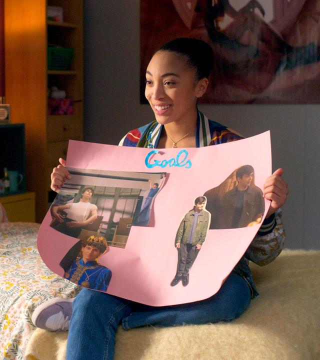 Cece sitzt lächelnd auf ihrem Bett und hält ein selbstgemachtes Plakat in die Luft, auf dem sie Bilder von Adrian und Benoit gepostet hat.