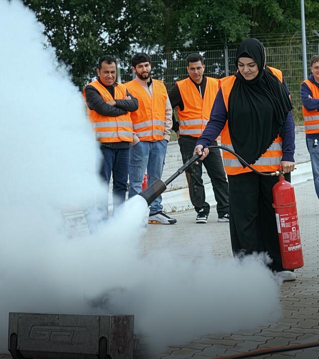 Eine Frau mit Warnweste löscht ein Feuer mit einem Feuerlöscher bei einer Brandschutzübung. Im Hintergrund stehen weitere Teilnehmende in Warnwesten und schauen zu. Über dem gelöschten Feuer ist weißer Rauch.