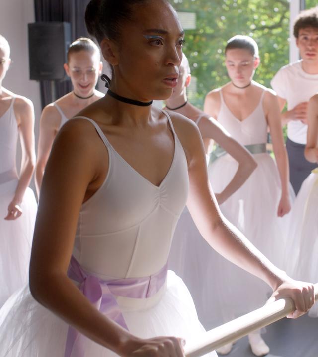 Cece steht in einem weißen Ballettkostüm angestrengt an der Balletstange und wird von ihren Mitschülern beobachtet.