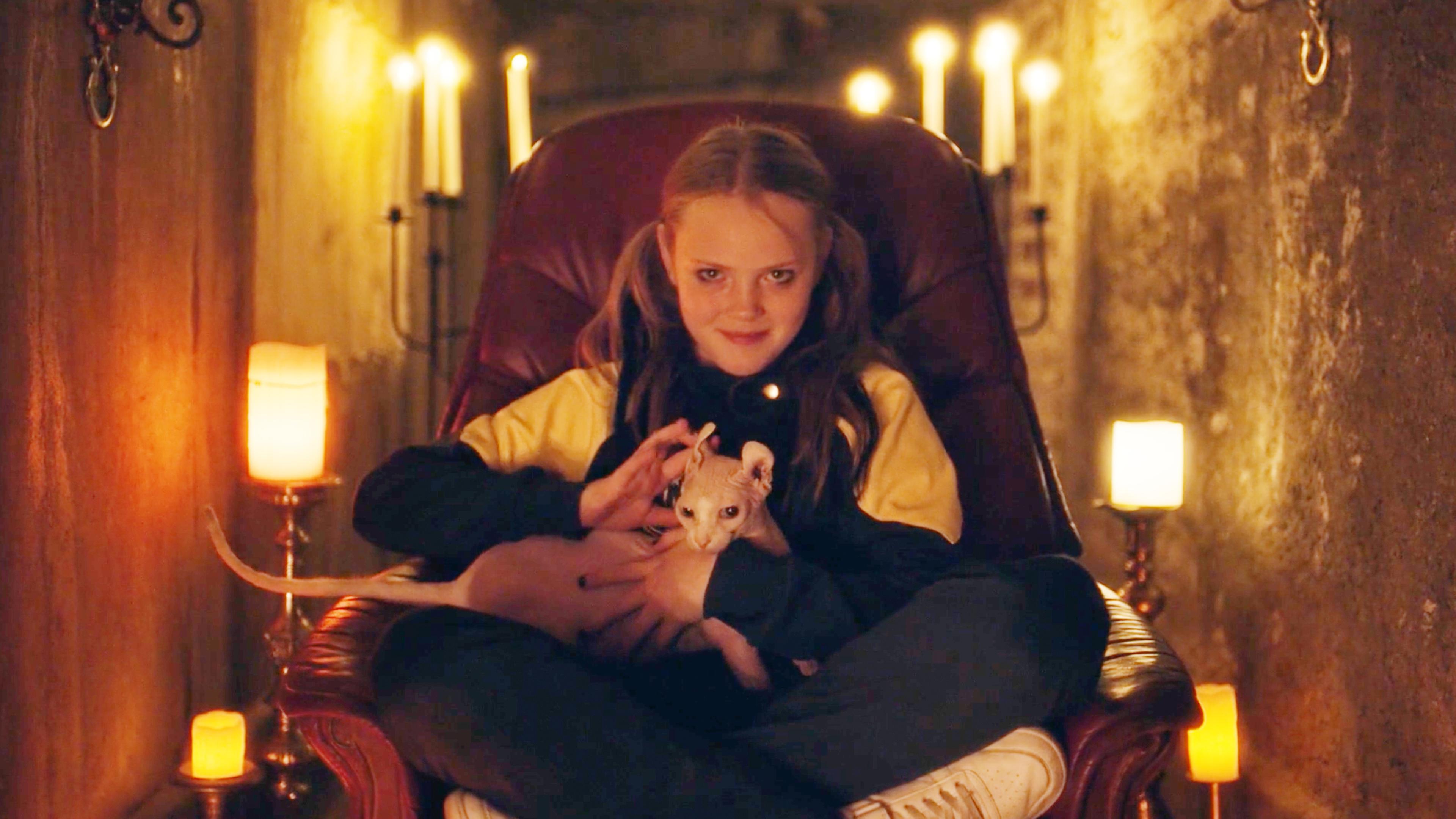 Im Keller der Schule: Tele sitzt fies grinsend im Schneidersitz auf einem Ledersessel. Auf ihrem Schoß sitzt eine Katze. Um sie herum sind kahles Mauerwerk und brennende Kerzen zu sehen.