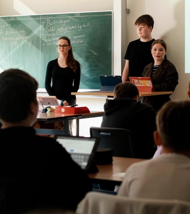 Jana (14) steht mit zwei Freunden an der Tafel vor einer Schulklasse mit jüngeren Schülerinnen und Schülern.