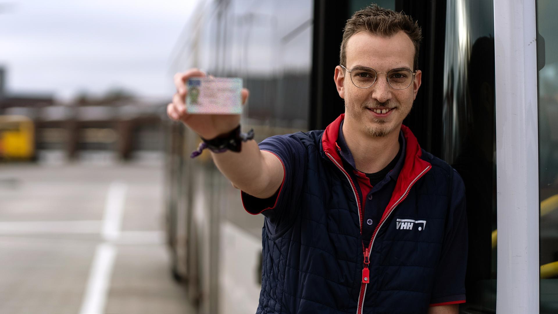 Ein junger Mann steht, mit einer Weste der Verkehrsbetriebe Hamburg-Holstein (vhh) bekleidet, neben einem Linienbus. In seiner rechten Hand hält er seinen Busführerschein in die Kamera. Niels kann damit in seinen neuen Job als Busfahrer starten.