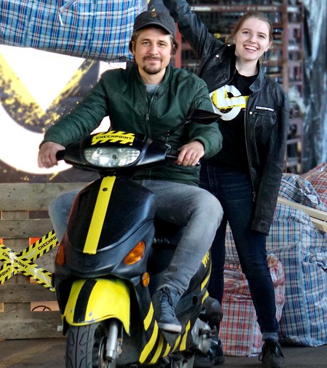 Checkpoint: Moderator Tommy Scheel sitzt auf einem Motorroller, während drei Mitglieder des Checkpoint-Teams um ihn stehen und das Checkpoint-Logo, sehr große Einkaufstaschen oder Autorreifen tragen. 